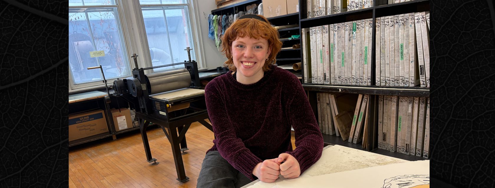 A person with red curly hair wearing a black headband and purple sweater sits on a stool leaning on a table with art prints smiling into the camera with a printing press, shelves and a window behind her.