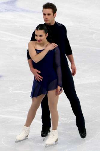 Two figure skaters stand posing in dark-coloured costumes on the ice.