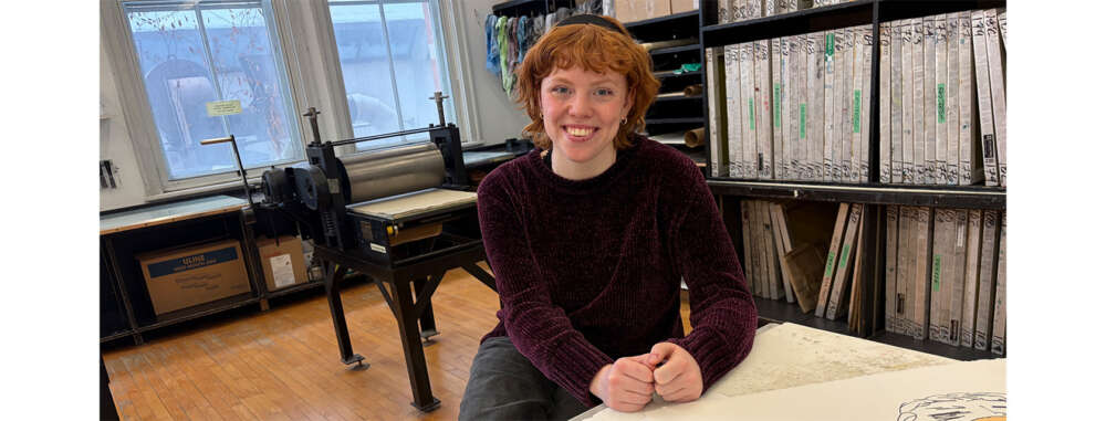 A person with red curly hair sits on a stool leaning on a table with art prints