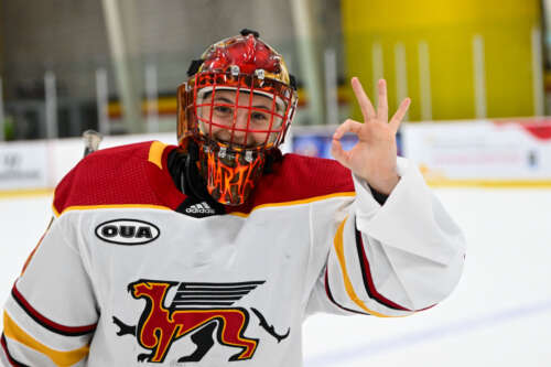 A Gryphon women's hockey goalie stands on the ice in her white, red and gold uniform flashing the okay sign with her hand while smiling at the camera.