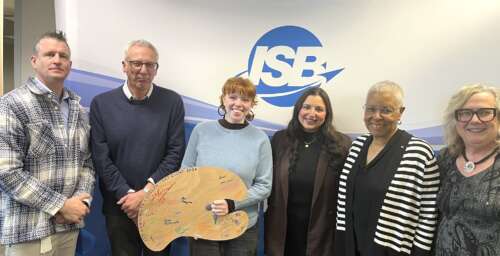 A group of six people stand in a row smiling against a white wall with a blue logo that reads ISB.