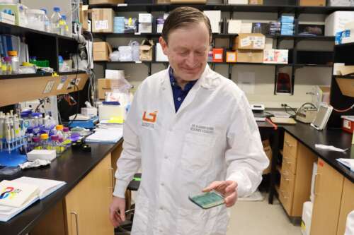 A researcher in a white lab coat stands holding a sample to the camera with cupboards and lab equipment in the background.