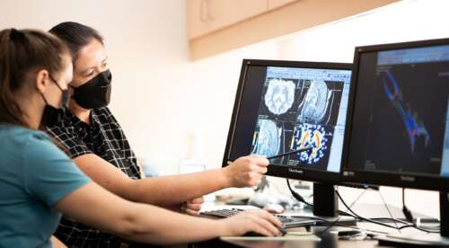 Two people wearing black face masks look at two computer screens showing medical imaging.
