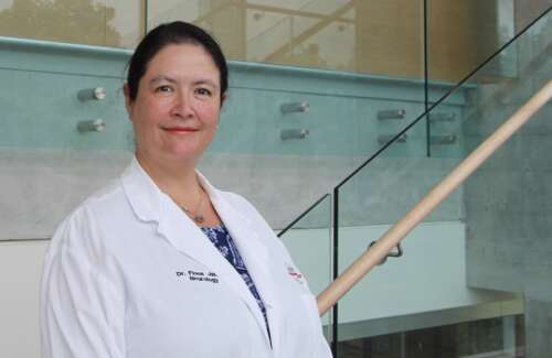 A person with brown hair pulled back wearing a white lab coat over a blue patterned top stands in front of a glass and wood staircase smiling into the camera.
