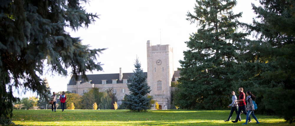 Students walk across green campus space known as Johnston Green in front of gray building Johnston Hall in the summer.
