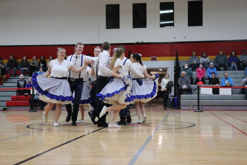 dancers in white dresses and suits perform on a brown gym floor