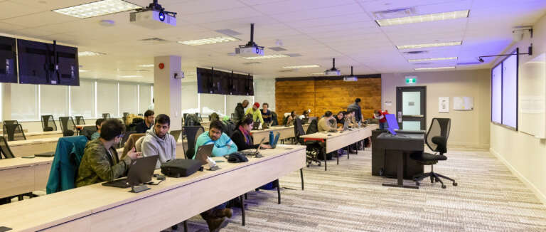 wide shot of a bright modern classroom with students at long desks on laptops