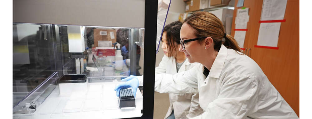 Two people in lab coats reach into a class enclosed machine to retrieve samples