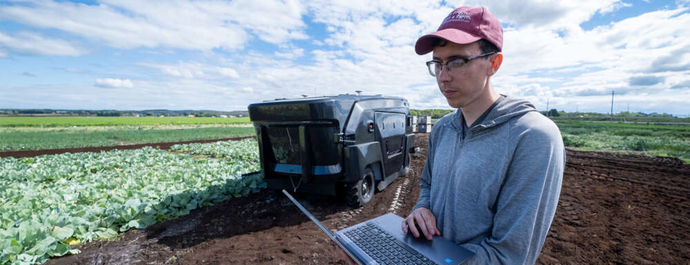 a person a red baseball cap, glasses and green zipped sweatshirt looks at a laptop computer in a field in front of self-driving farm equipment