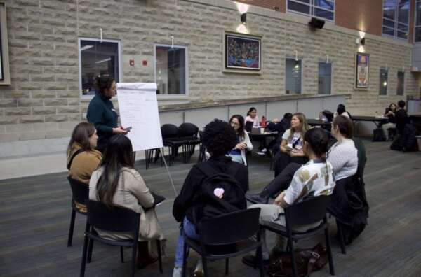 A group of people sit in a discussion circle. One person stands next to an easel with a notepad with notes.