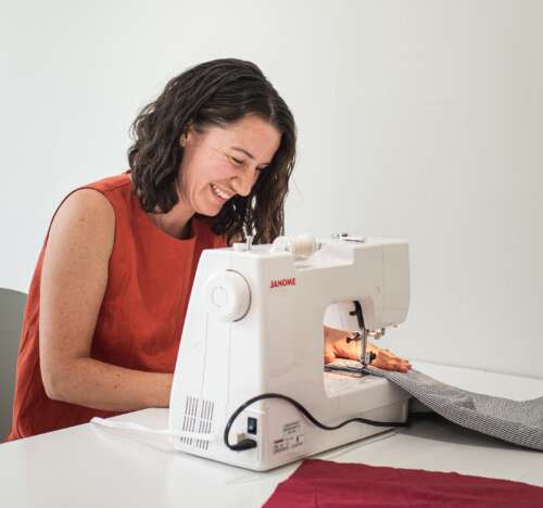 A person with curly brown hair wearing an orange dress smiles as they bend over a white sewing machine sewing grey fabric.