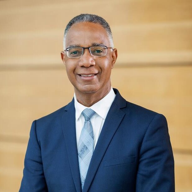 A man with black and grey hair wearing a blue suit and tie and wire frames smiles into the camera in front of a beige background