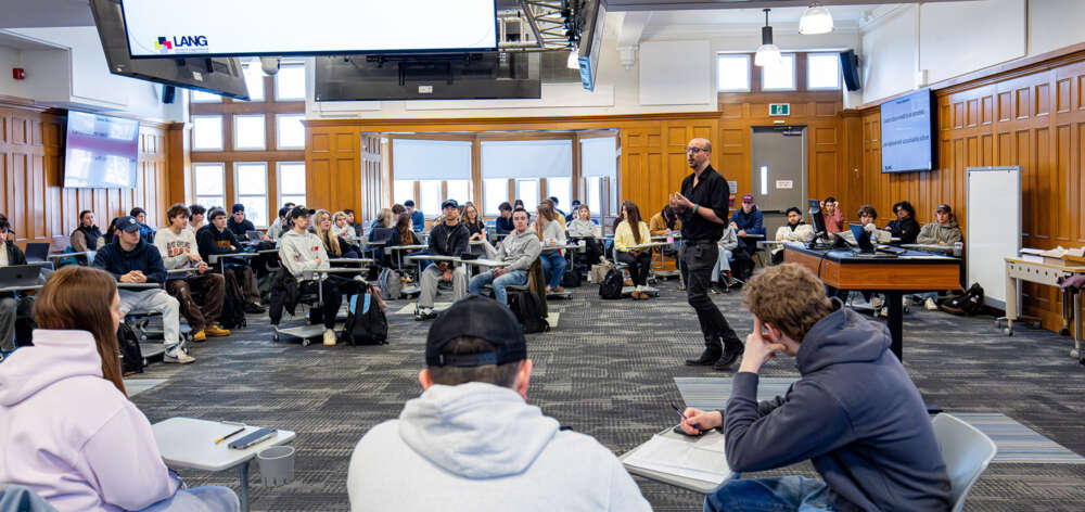 a wide view of a wood panelled classroom filled with students at desks with an instructor in the centre
