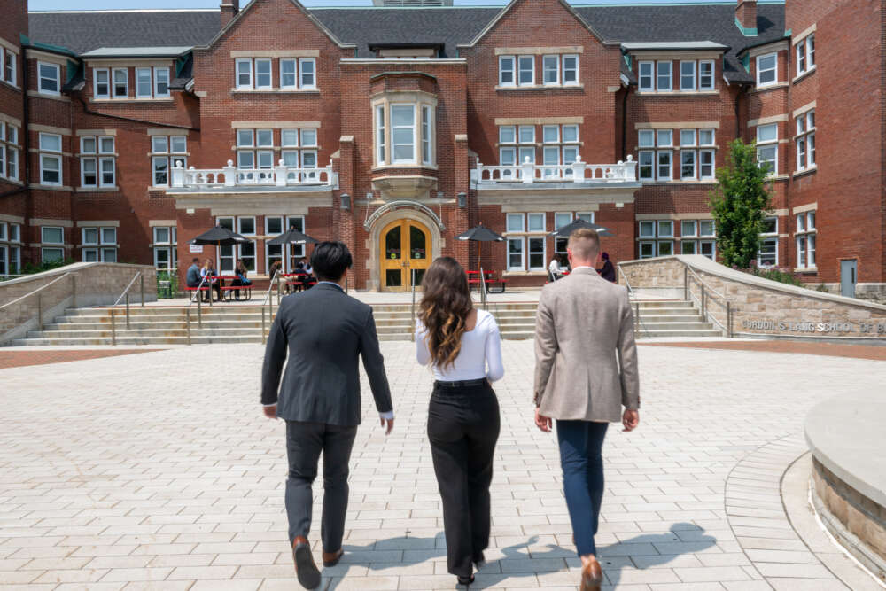 The backs of three students walking toward the Lang School of Business red brick building on a sunny day