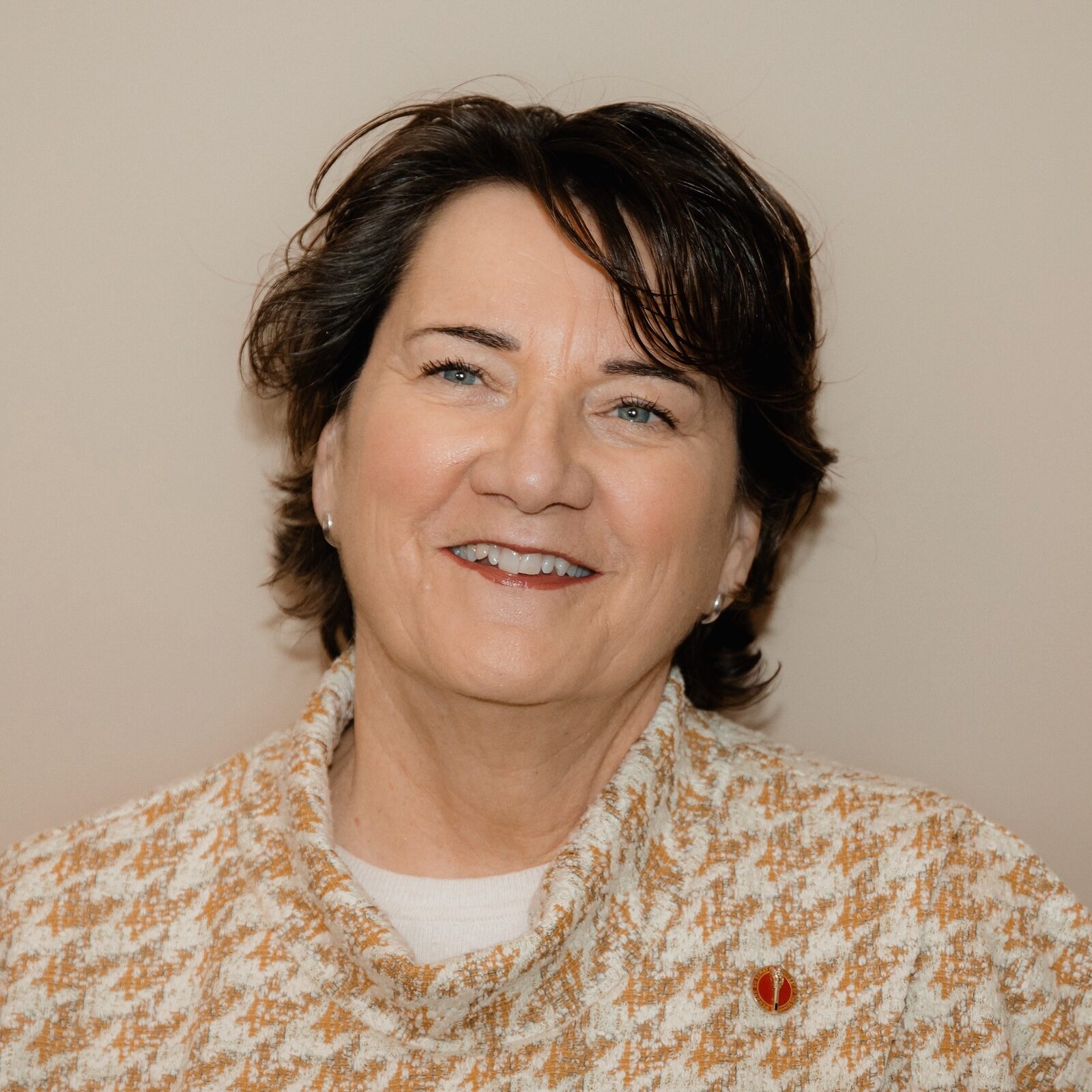 A person with short brown hair wearing a beige and white houndstooth jacket smiles into the camera in front of a beige background.