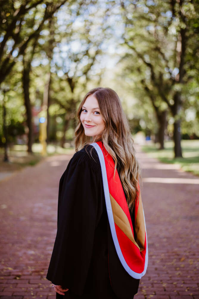 person in black grad gown and red hood poses on brick path