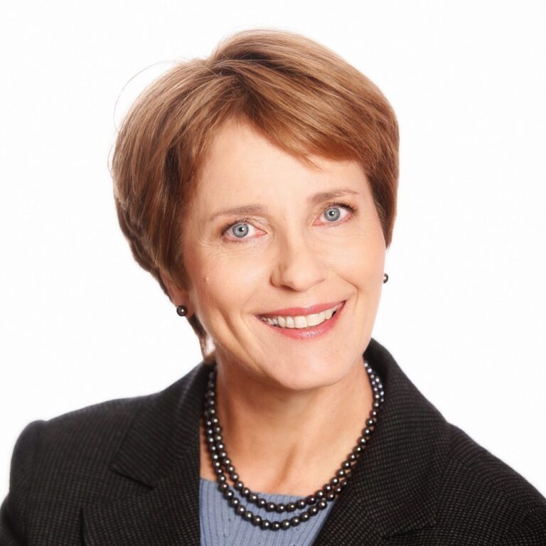 A person with short brown hair wearing a light blue shirt under a black blazer with a black bead necklace smiles into the camera in front of a white background.
