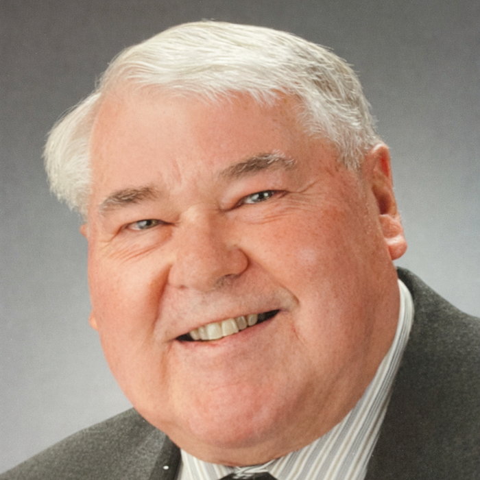 A person with white hair wearing a charcoal suit, striped shirt and dark tie smiles into the camera in front of a grey backround.