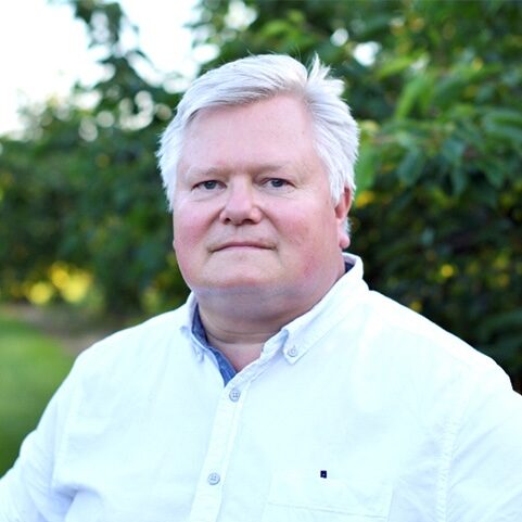 A man with white hair wearing a white buttoned shirt stands in front of greenery outside smiling into the camera.