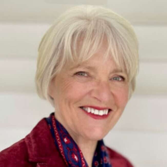 A person with short grey hair wearing a red jacket and patterned scarf smiles into the camera in front of an off-white background.