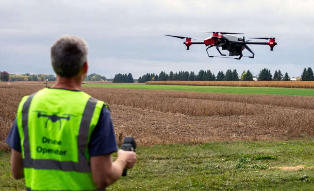 The back of a man in a yellow vest operating a large drone over a new crop field