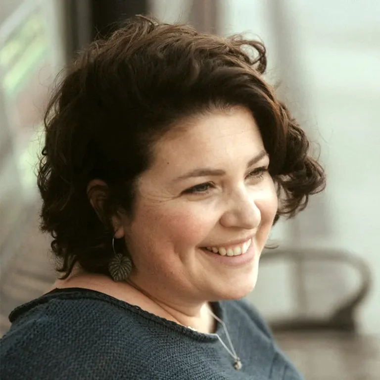 A person with short brown curly hair wearing a black top is photographed from their right side, smiling into the distance.