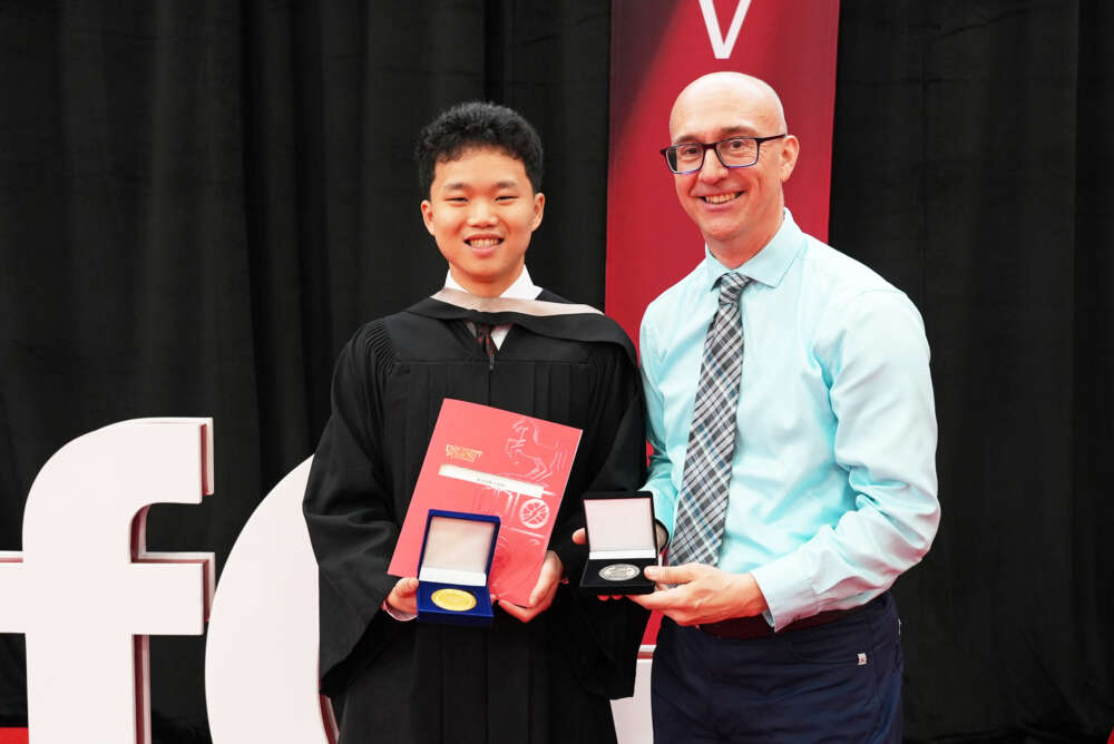 A student in convocation regalia holds a degree and a medal while a man in a tie stands beside him holding another medal