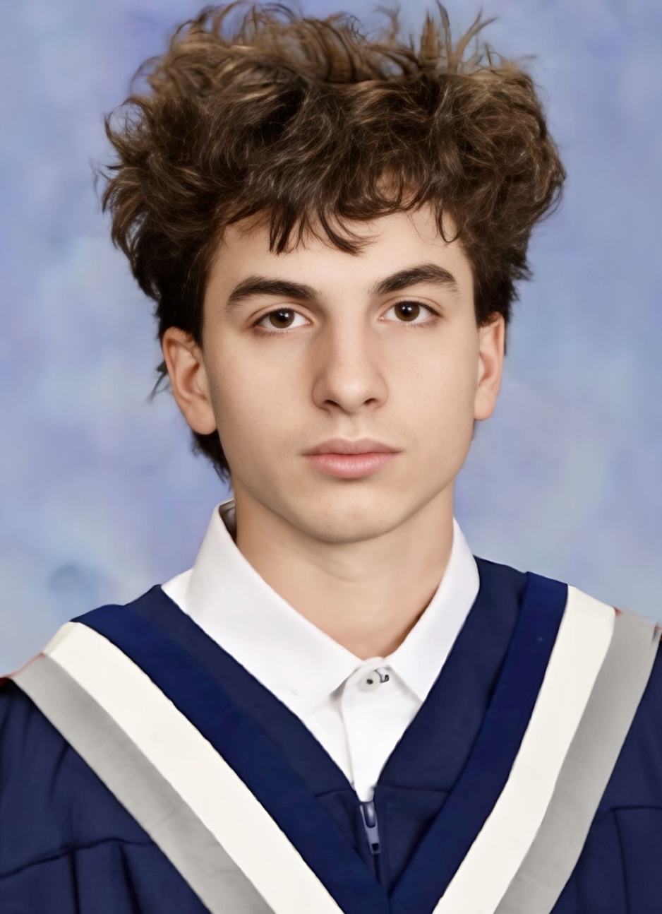 A student with short brown hair wearing a white button-up shirt under a blue graduation gown with a blue, white and grey collar looks into the camera in front of a blue marble background.