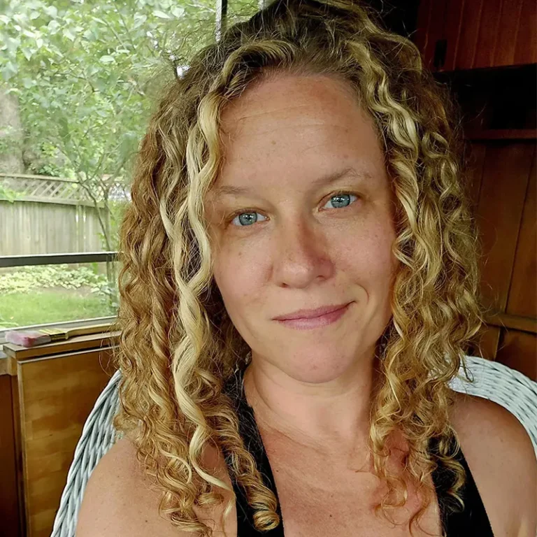 A person with blonde curly hair wearing a black tank top smiles into the camera, sitting in a white rattan chair in front of a window with trees in the background.