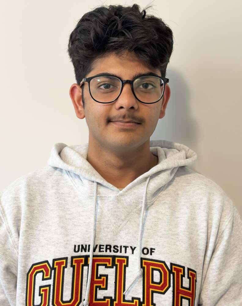 A student with short brown hair and black frame glasses wearing a grey U of G hoodie stands in front of a cream background looking into the camera.