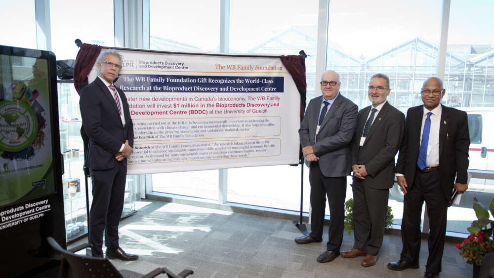 Four people in suits stand next to a sign announcing a gift for the BDDC