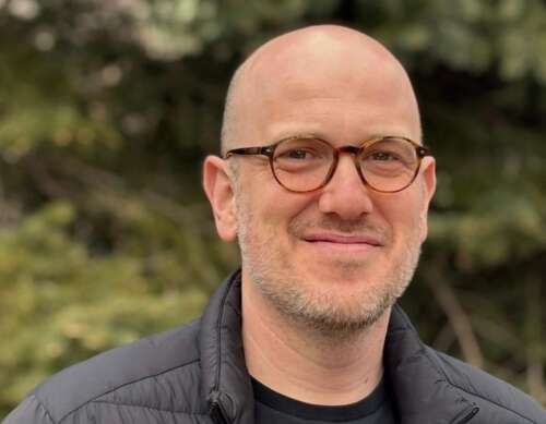 A bald person wearing brown glasses and a charcoal puffer jacket stands smiling in front of an evergreen tree