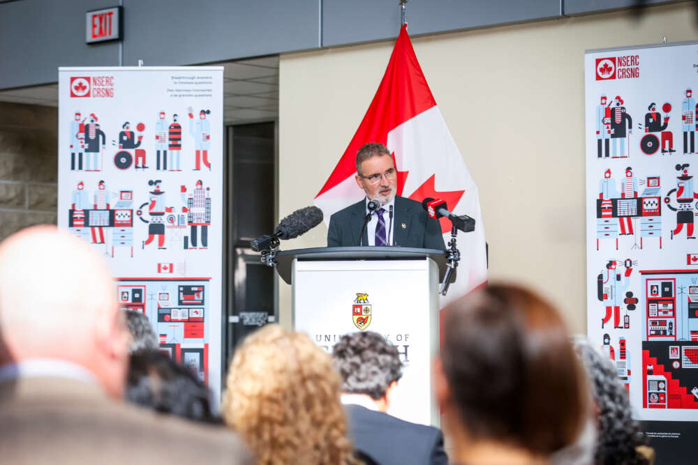 person in suit stands at podium in front of red and white flag