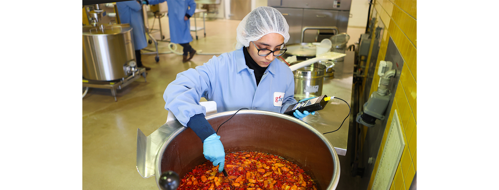 person in lab coat and hairnet holds a sensor near a vat filled with peppers and liquid