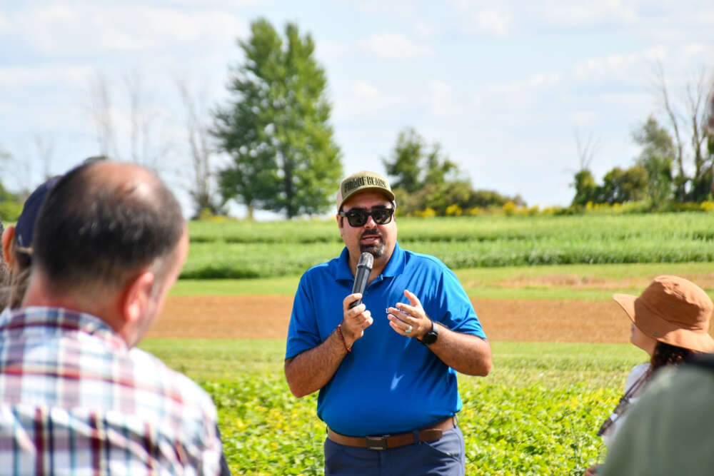 person in blue shirt speaks in front of a crowd with a field in the background