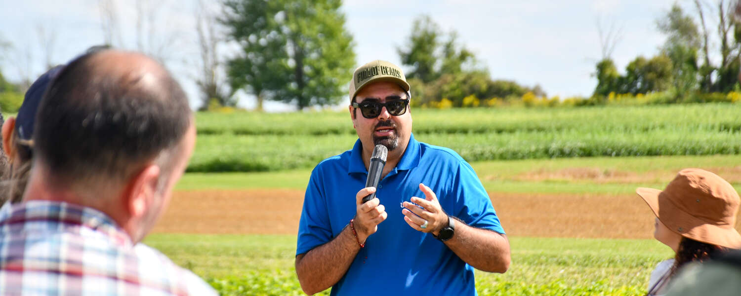 person in blue shirt speaks in front of a crowd with a field in the background