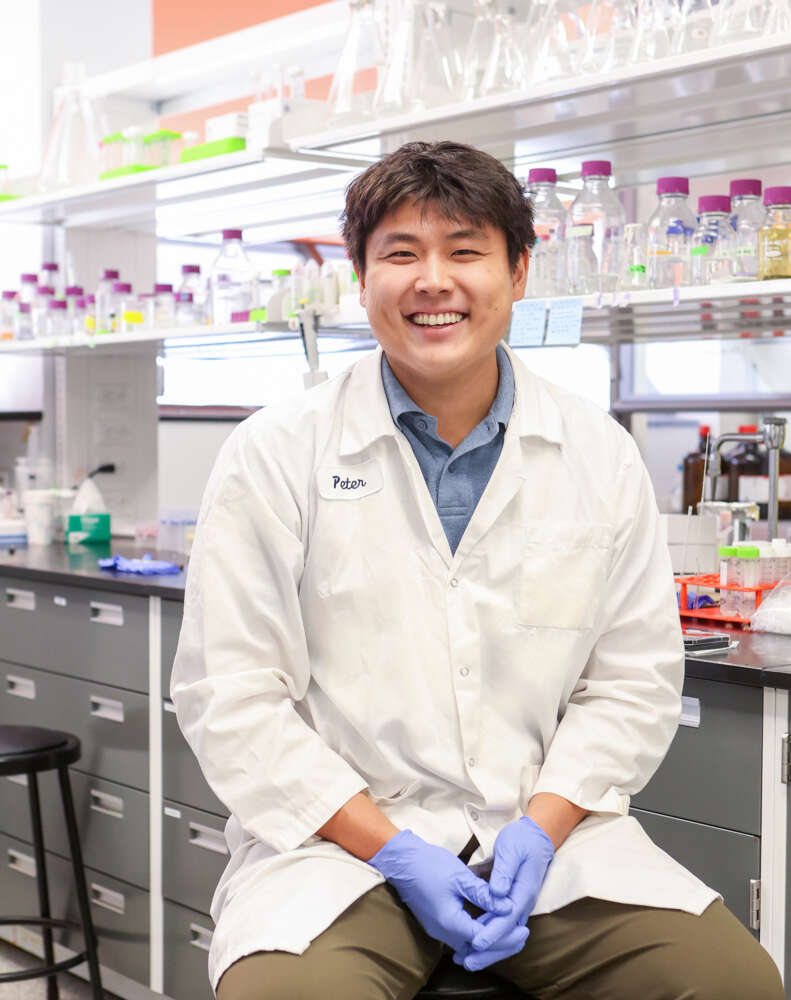 person in a lab coat smiles while sitting on a stool in a chem lab