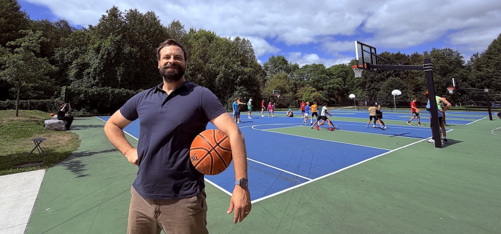 A man with brown hair and a beard wearing a blue polo shirt and brown pants holds a basketball under one arm while people play basketball on a blue and green court behind him.