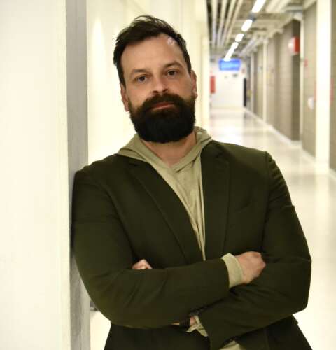A person with short brown hair and a brown beard wearing a cream hoodie under a dark blazer stands against a white wall with arms crossed, staring into the camera.