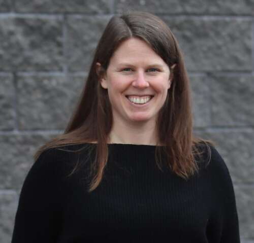 A person with long brown hair wearing a black sweater stands smiling at an angle in front of grey brick.