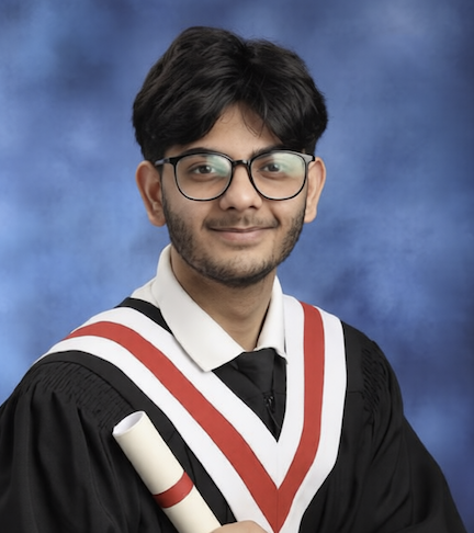 A student with brownhair and a beard wearing black framed glasses smiles wearing a black graduation robe holding a scroll in front of a blue background.