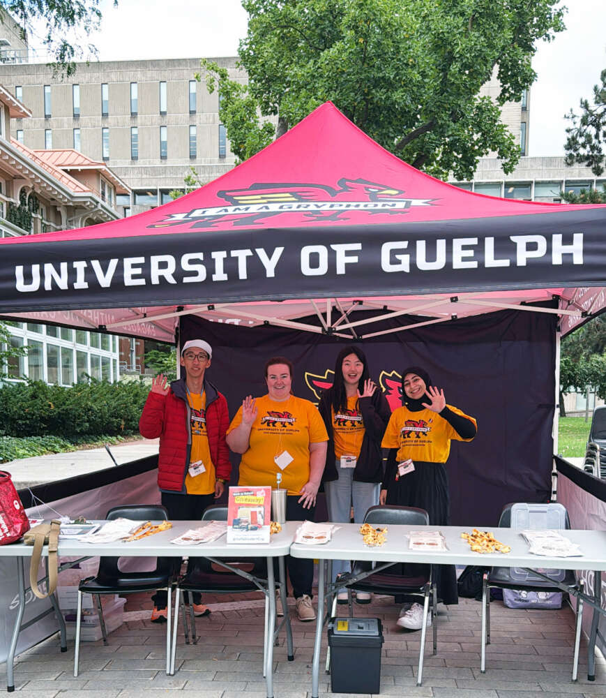 Four people in yellow U of G t-shirts wave to the camera while under a U of G folding tent
