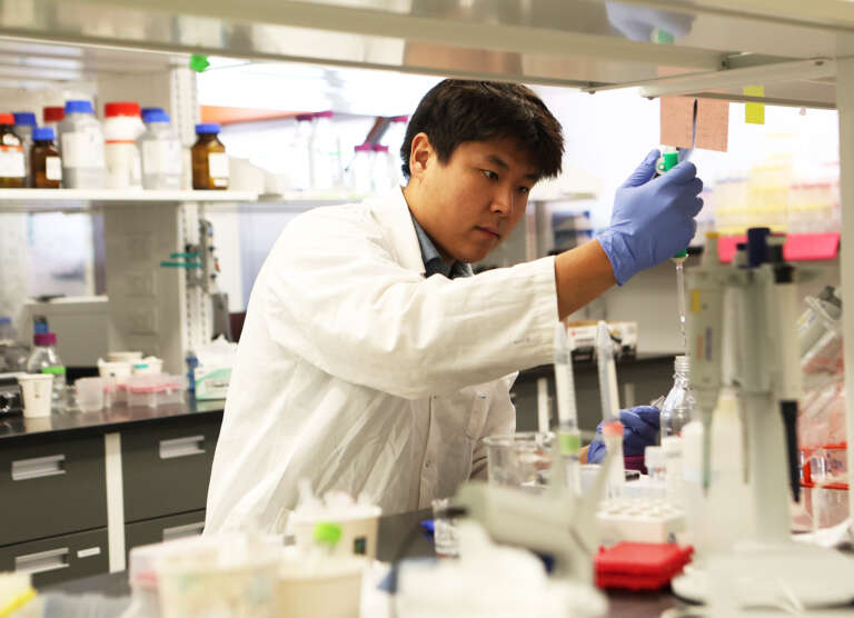 person in a lab coat uses a long pipette to extract a clear liquid in a chemistry lab