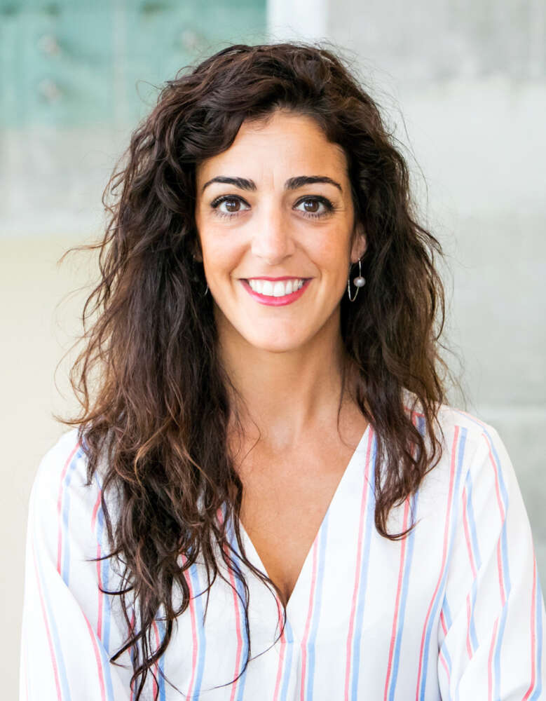 A person with long brown curly hair wearing a white blue and red striped blouse smiles into the camera in a bright atrium.