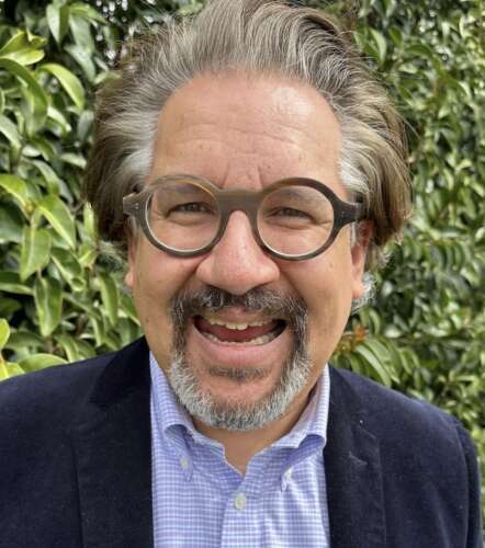 A person with brown and gray hair and beard wearing brown, round frame glasses, a blue button-up shirt and blue blazer smiles in front of a wall of greenery.