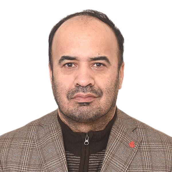 A person with short brown hair wearing a brown plaid blazer over a brown striped sweater with a zipper looks into the camera in front of a white background.