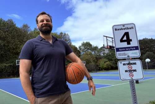 A person with brown hair and a brown beard wearing a navy polo shirt stands in front of a basketball court beside a city sign that reads Norm Jary Park