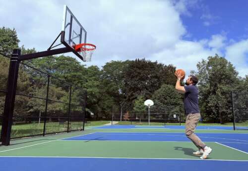 A person with brown hair and a brown beard wearing a navy polo shirt and light brown pants stands mid-shot on a blue and green basketball court surrounded by trees.