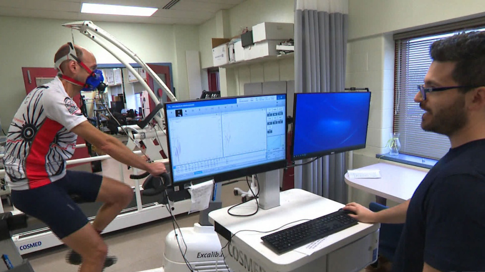 a person in cycling gear wearing an oxygen mask cycles while a second person evaluates results on a computer monitor