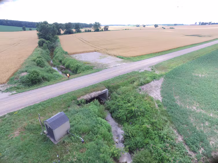 Aerial view of a creek running between 2 farm fields and under a gravel road